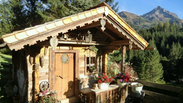 A wooden cabin with a porch, surrounded by greenery and mountains in the background.