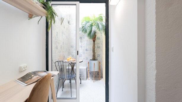 Une petite pièce comprenant un bureau, une chaise et une porte-fenêtre coulissante donnant sur un balcon où se trouvent un pot de fleurs et une petite table.