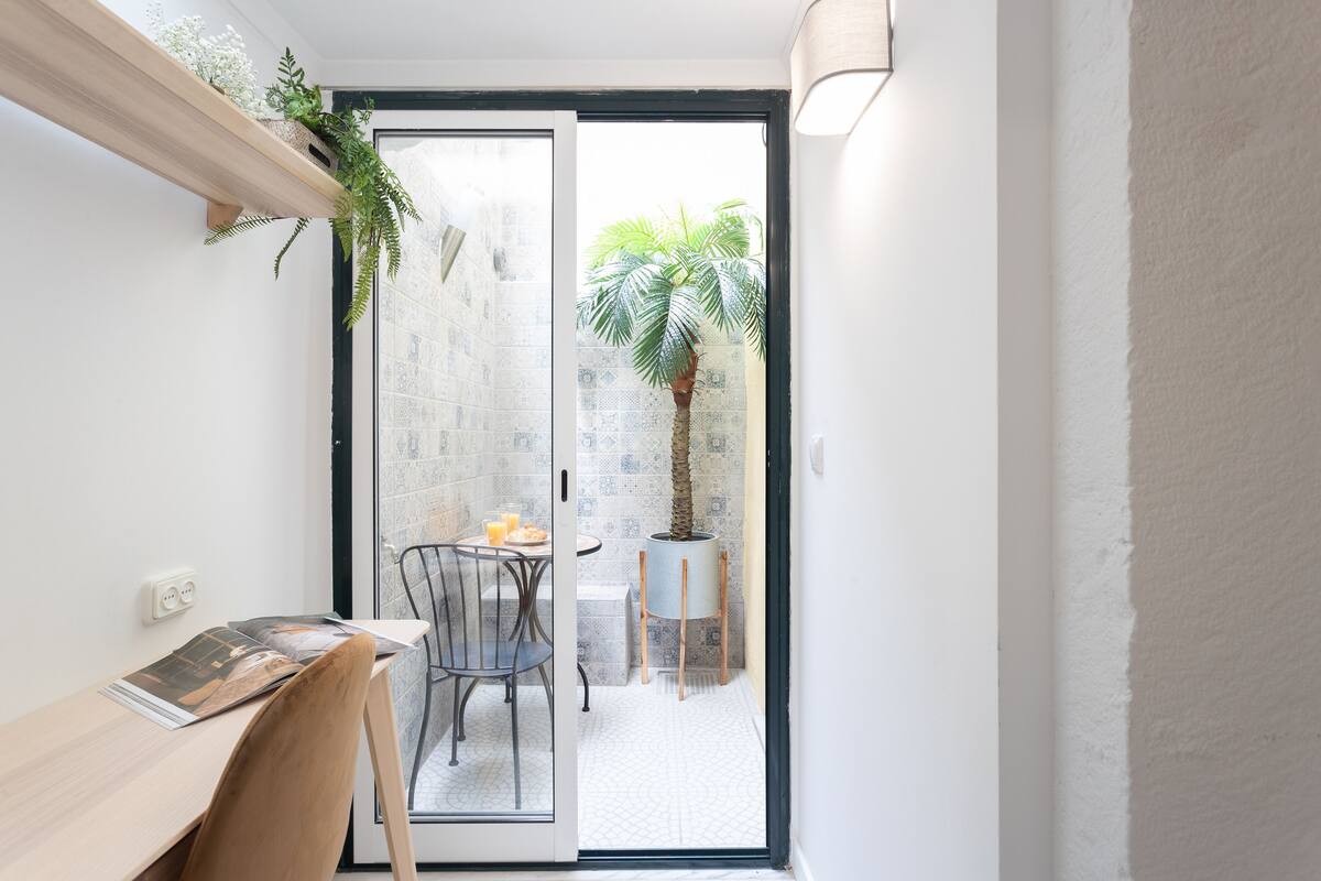 Una habitación pequeña con escritorio, silla y puerta de vidrio corredera que da a un balcón con planta en maceta y una pequeña mesa.