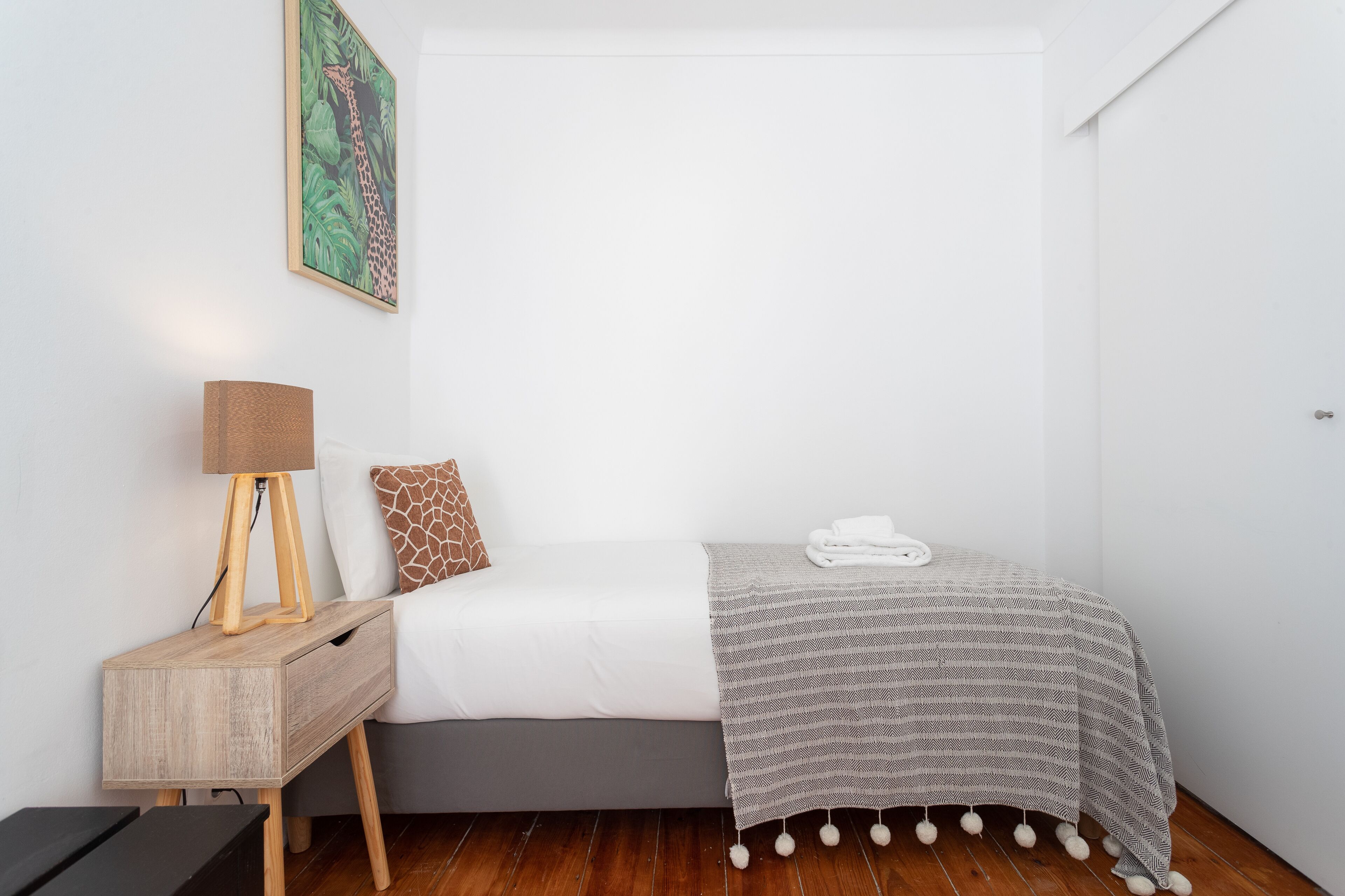 A neatly arranged bedroom with a bed, a bedside table with a lamp, a wall-mounted painting, and a neatly folded towel on the bed.