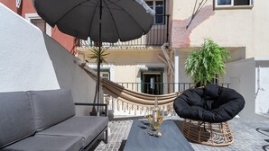 Une terrasse avec un canapé gris, une table avec une bouteille et des verres, un pouf noir et un hamac.