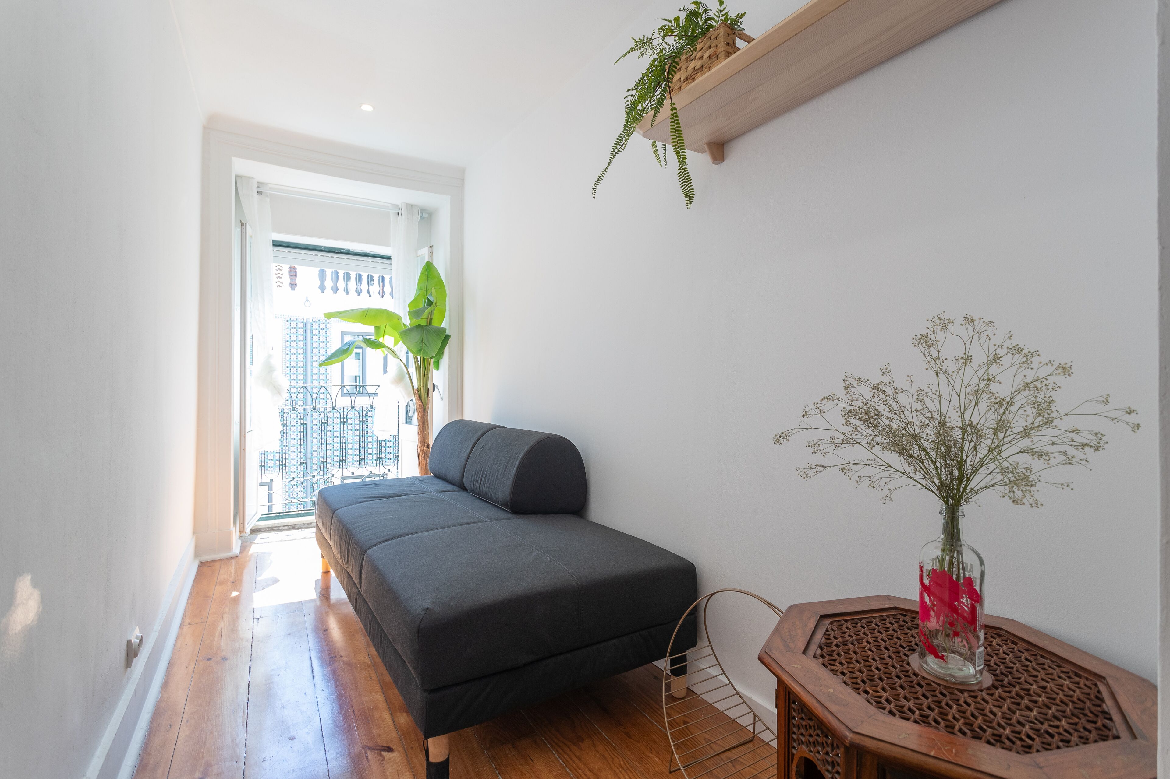 A small, modern bedroom with a bed, a wooden floor, a window with a view of buildings, a potted plant, and a decorative vase.