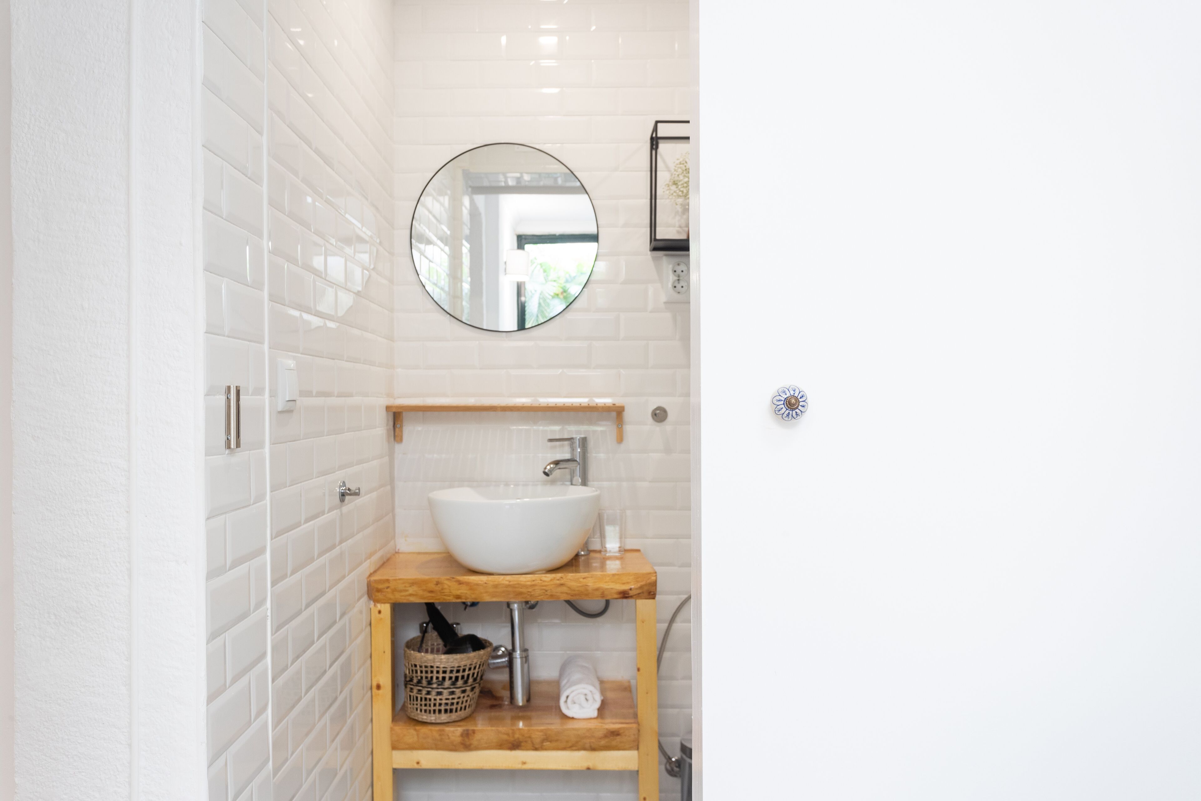 Een badkamer met een witte betegelde muur, een houten wastafelmeubel met een wastafel, een ronde spiegel en een lijst met een foto aan de muur.