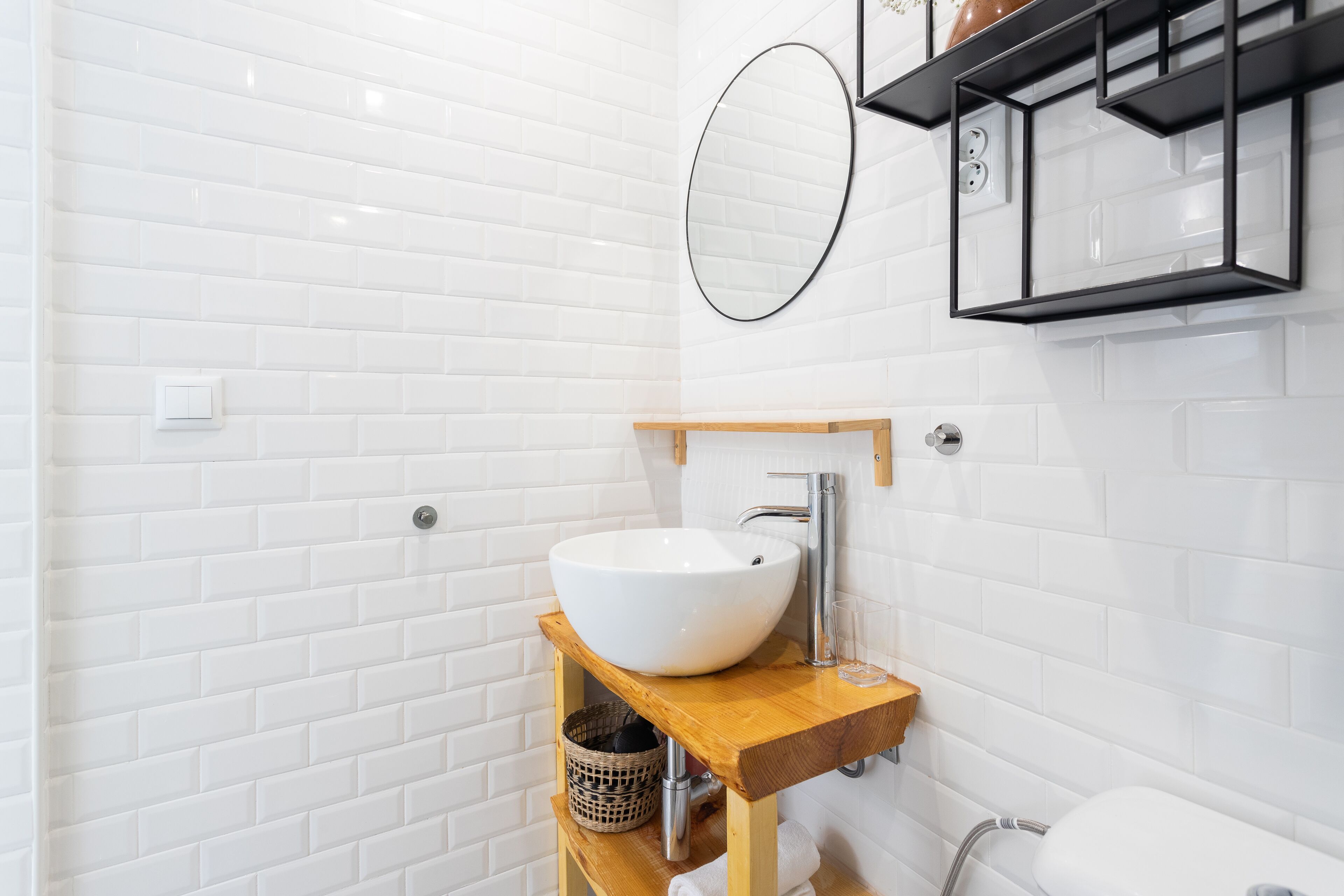 A bathroom with a white tiled wall, a wooden vanity with a white sink, a mirror, and a shelf with toiletries.