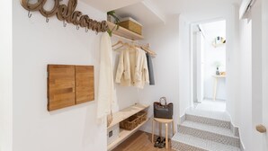 Un couloir avec une armoire en bois, un porte-manteau avec des vêtements, un panier en osier, un petit tabouret sur lequel est posé un sac à main, et un escalier dont les marches sont recouvertes de carreaux décoratifs.