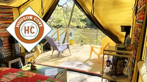 A tented accommodation with a view of a river and trees, a wooden deck, a table with a menu, and a bed with a floral bedspread.