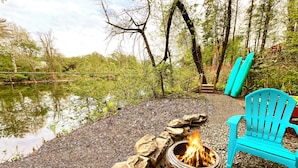 A campfire with rocks, a blue chair, and a wooden deck overlooking a body of water.