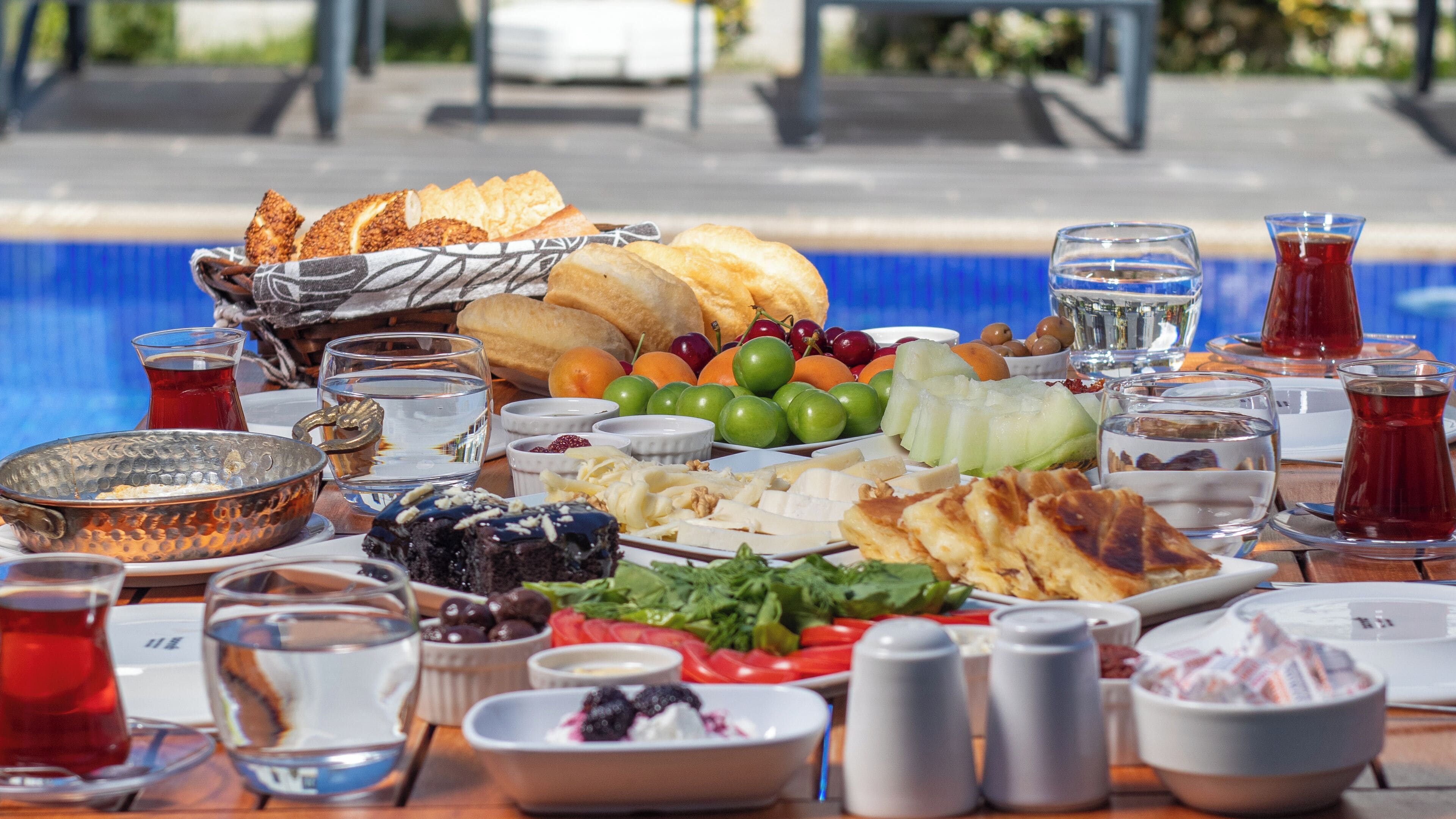 A table with a variety of food items and drinks, including bread, fruits, cheese, and olives, set outdoors with a pool in the background.