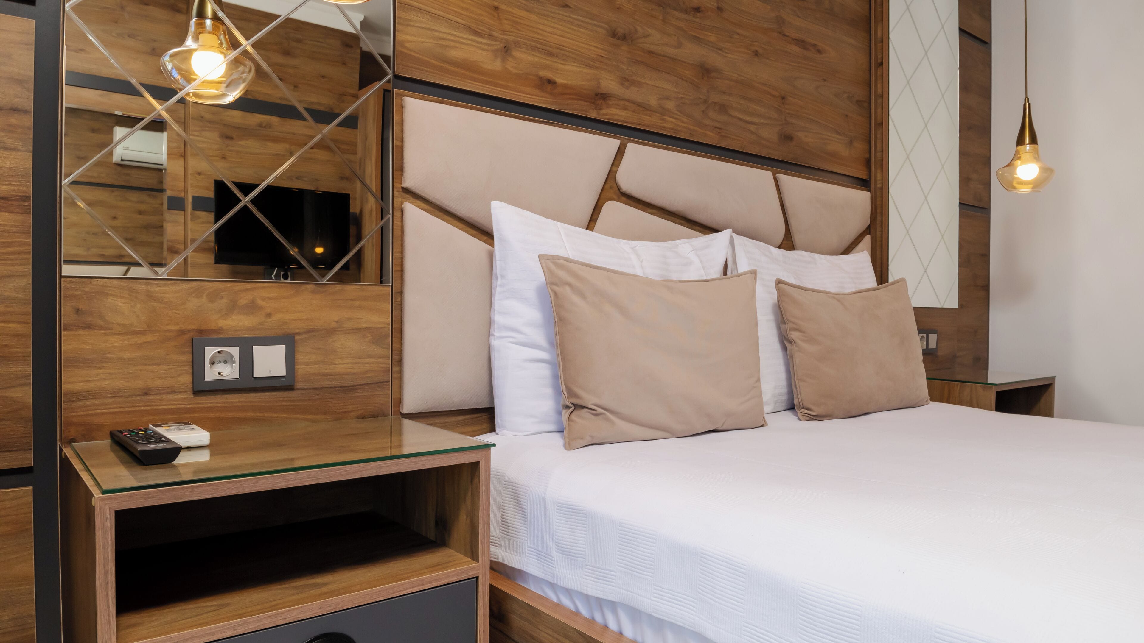 Ett modernt hotellrum med en sänggavel i trä, ett sidobord i glas, en fjärrkontroll och en väggmonterad luftkonditioneringsanläggning.