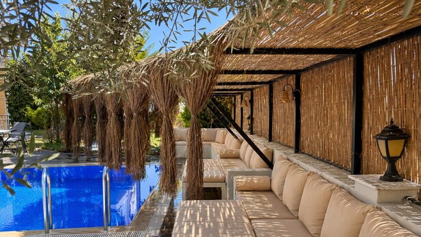 A poolside area with a blue pool, beige cushioned seating, and a thatched roof structure. - Cunda Garden  (Ayvalik)