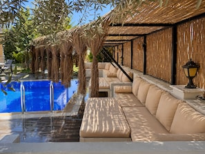 A poolside area with a blue pool, beige cushioned seating, and a thatched roof structure.