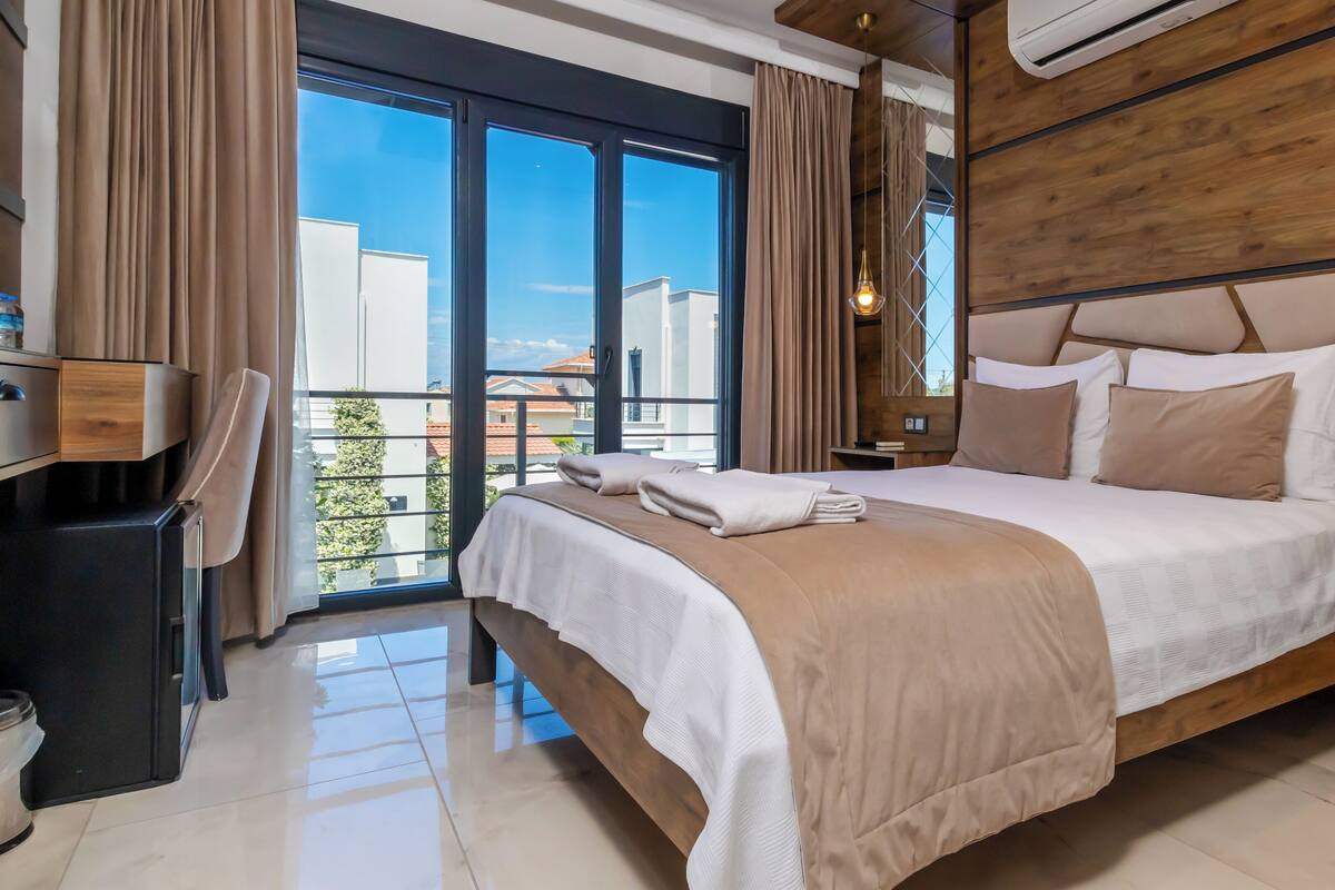 Ein modernes Hotelzimmer mit einem großen Bett, einem Holz-Kopfteil und Blick auf die Stadt durch raumhohe Fenster.