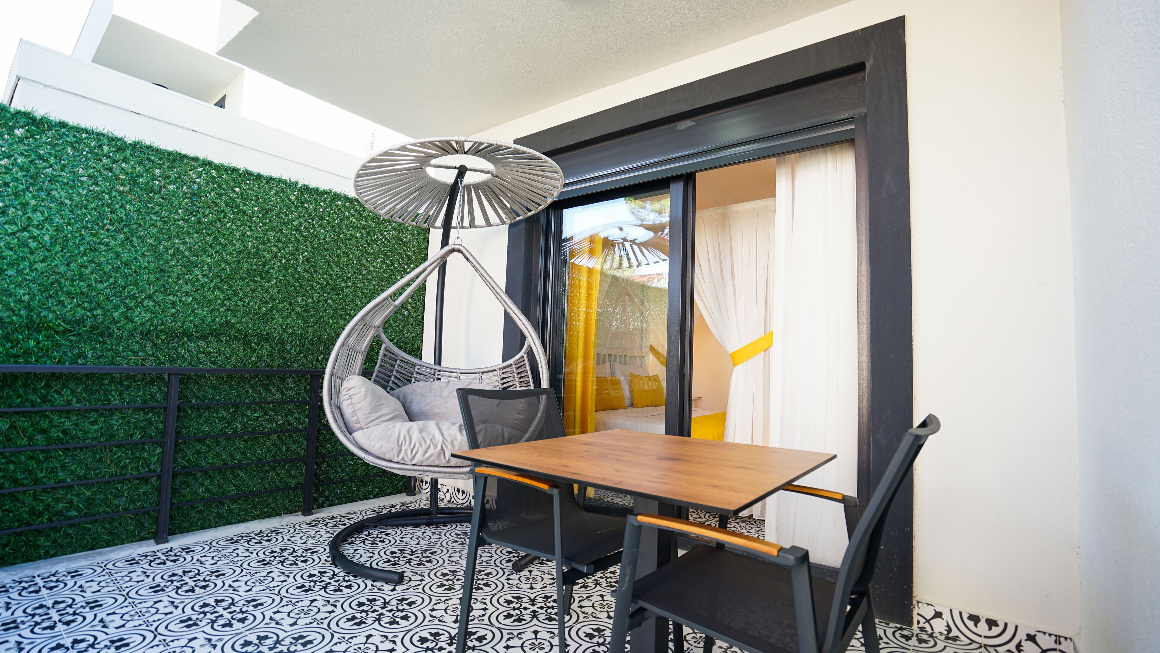 A balcony with a hanging chair, a wooden table, and a patterned floor.