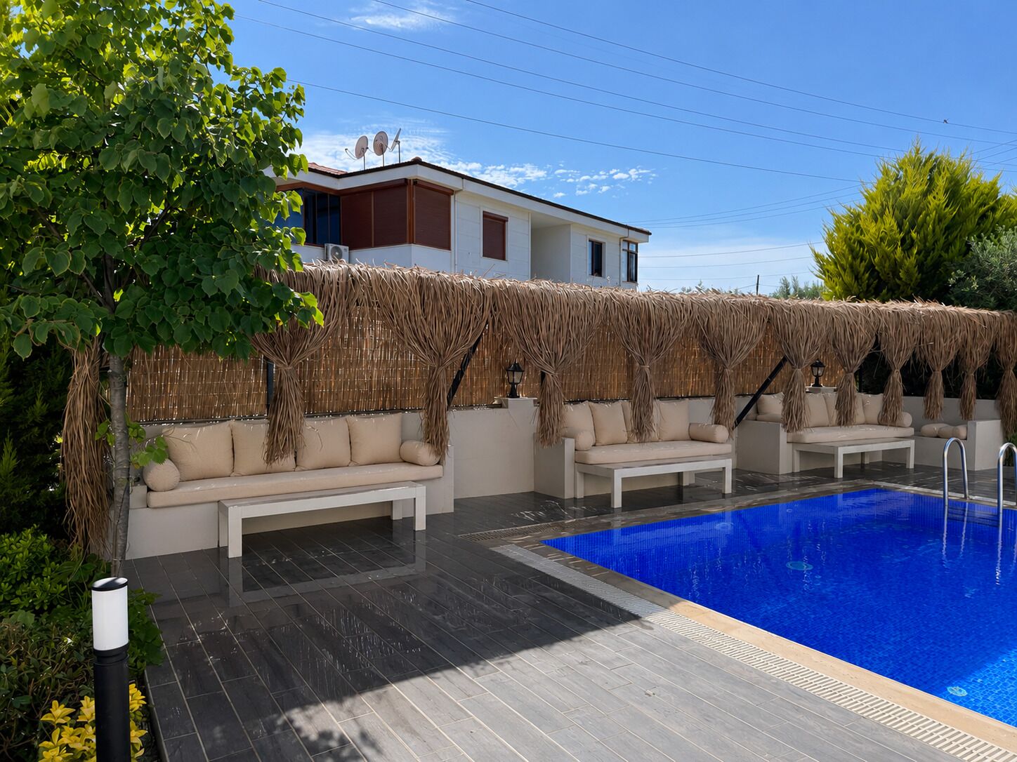 Un área junto a la piscina con piscina azul, sillas reclinables blancas y una estructura de techo de paja.