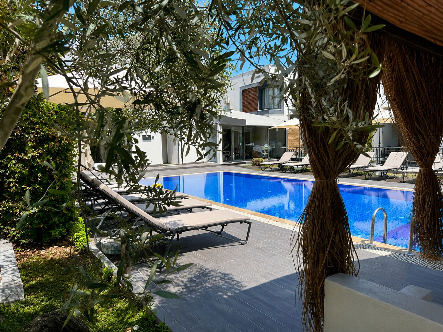 Un área de piscina moderna con sillones reclinables, piscina azul cristalina y un edificio con grandes ventanales.
