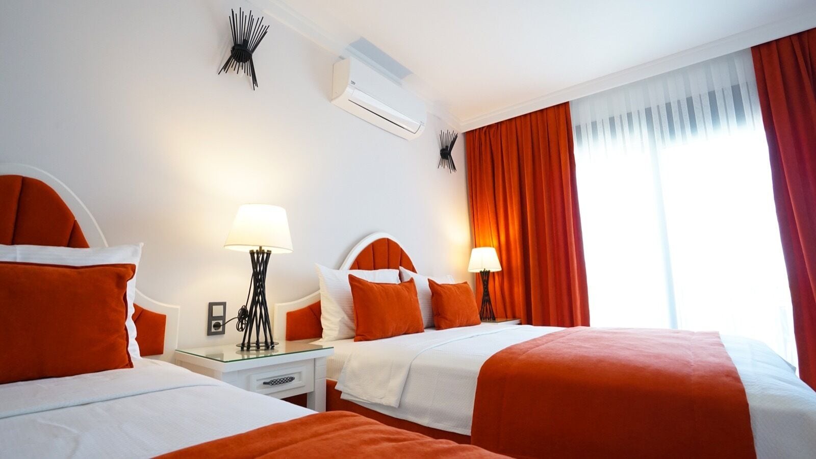 Ein Hotelzimmer mit zwei Betten, weißer Bettwäsche, orangefarbenen Akzenten, einem Nachttisch mit Lampe, einer Wand-Klimaanlage und einem Fenster mit roten Vorhängen.
