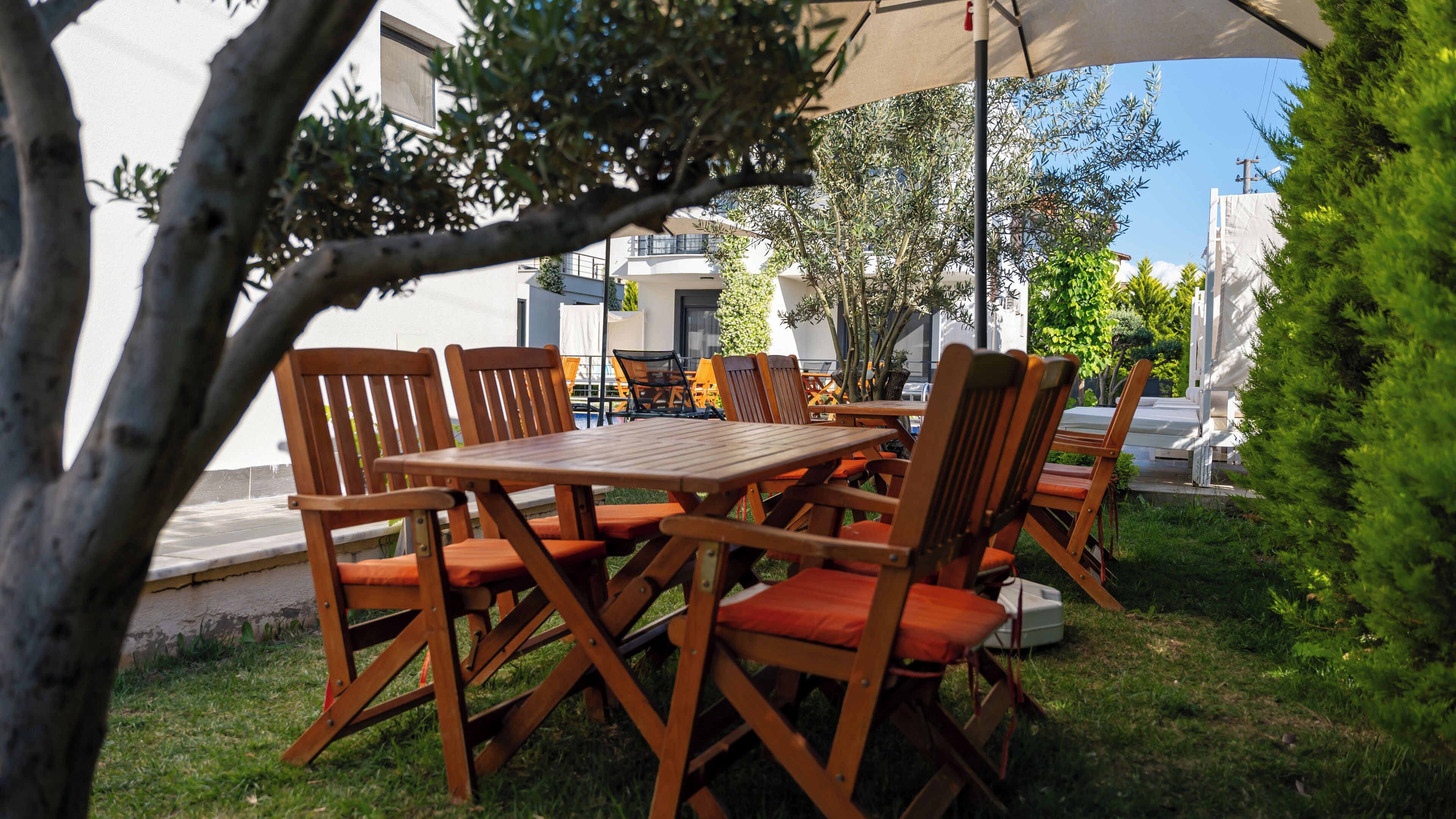An outdoor dining area with wooden tables and chairs, surrounded by greenery and a white building in the background.
