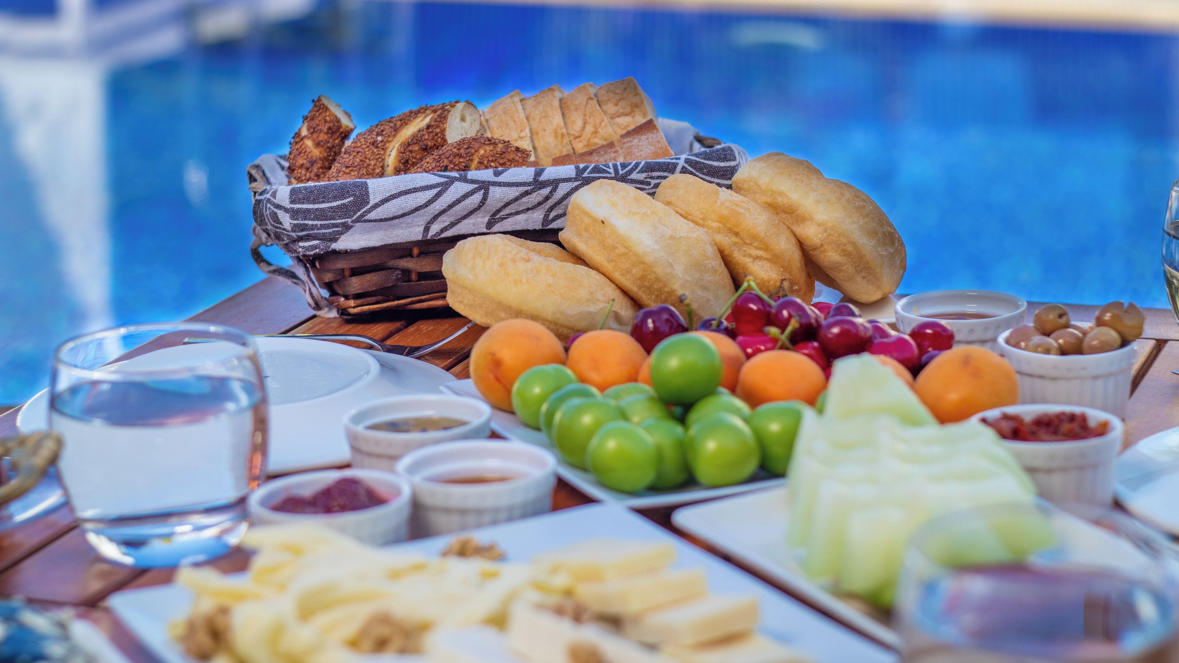 Une table garnie de divers aliments, notamment du pain, des fruits et des trempettes, est placée au bord d’une piscine.