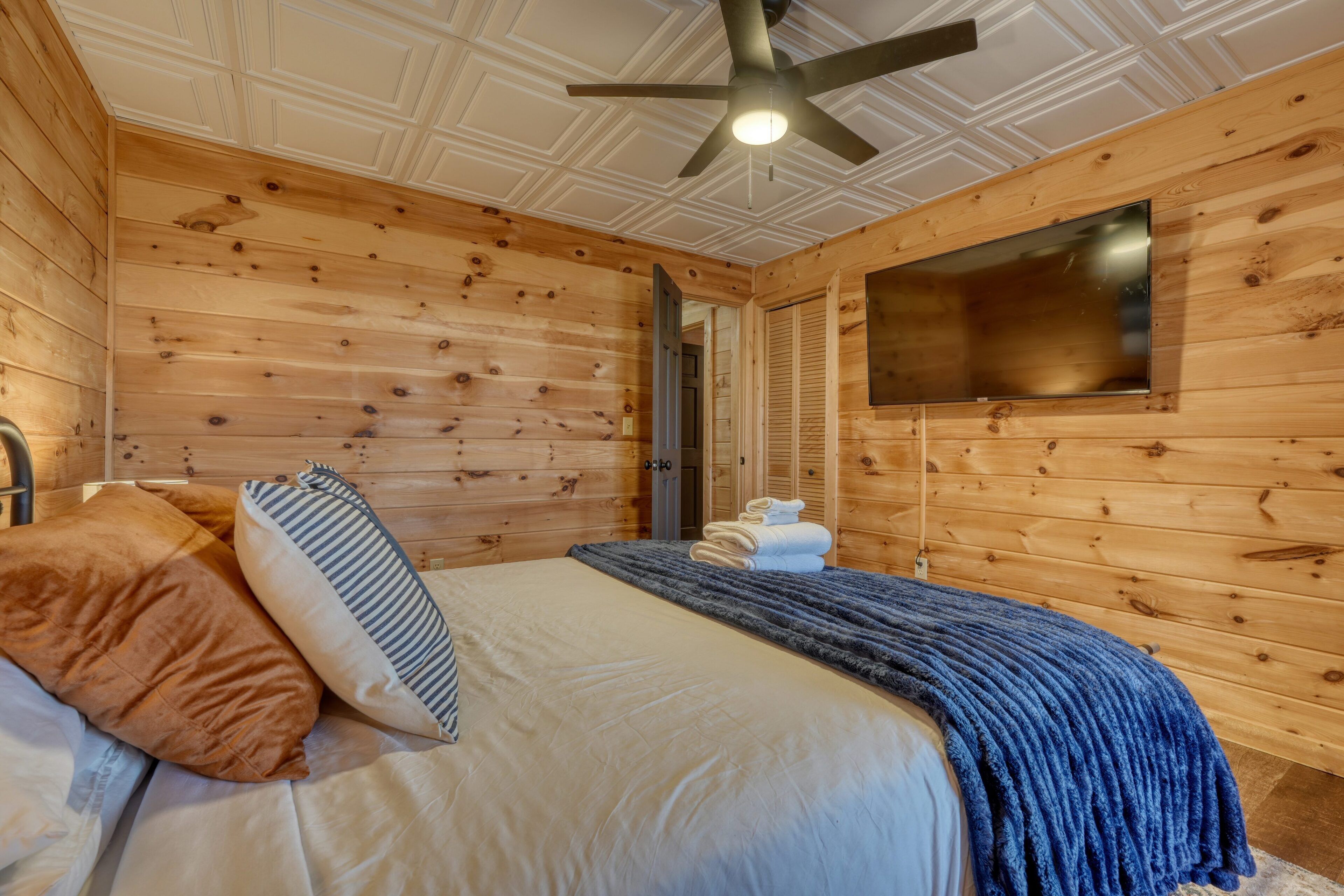 A wooden-paneled room with a bed, a ceiling fan, and a flat-screen TV mounted on the wall.