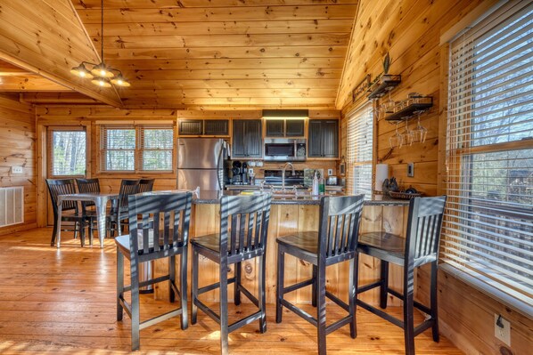 A wooden cabin interior with a kitchen area, dining table, and chairs. - Hidden Hills Sunset Soaks & Pizza Nights (Blairsville)
