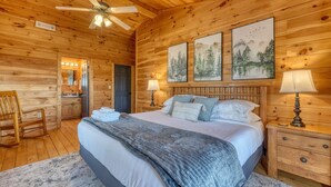 A wooden cabin bedroom with a bed, two bedside lamps, a ceiling fan, and framed artwork on the wall. - Hidden Hills, Cabin for Family Getaway with Mountain Views! - Hot Tub and Wifi (Blairsville)