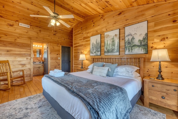 A wooden cabin bedroom with a bed, two bedside lamps, a ceiling fan, and framed artwork on the wall. - Hidden Hills Sunset Soaks & Pizza Nights (Blairsville)