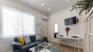 Un salon moderne comprenant un canapé, une table basse en verre, un bureau avec une chaise, un téléviseur fixé au mur et une plante en pot.
