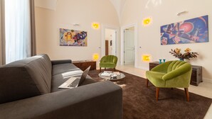 Ein modernes Wohnzimmer mit einem grauen Sofa, einem grünen Sessel und einem Couchtisch. An den Wänden hängen zwei Gemälde und eine Tür führt in einen anderen Raum.