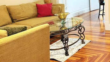 A living room with a sofa, a glass coffee table, and a patterned rug on a wooden floor.