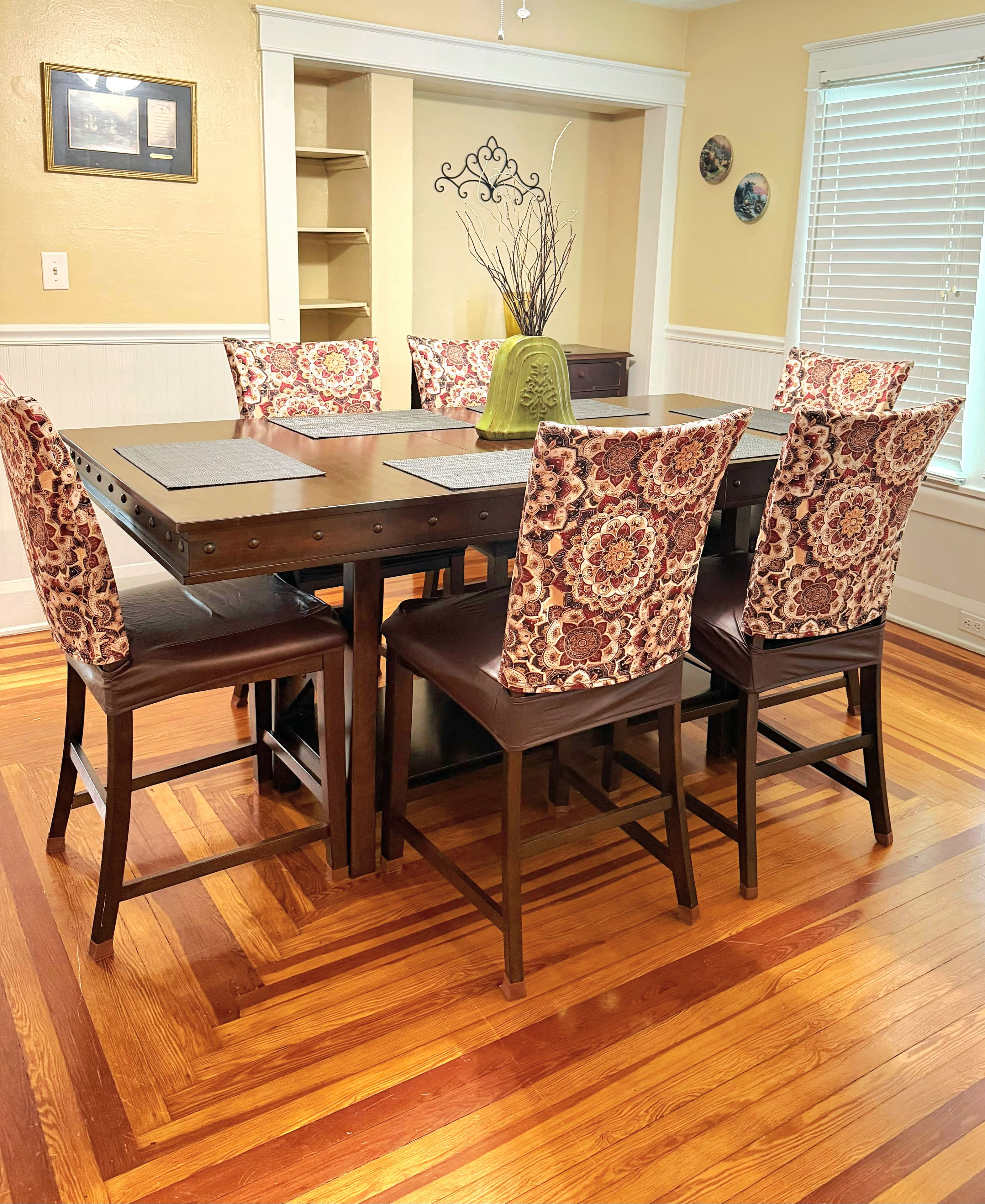 A dining room with a wooden table and patterned chairs, a vase with branches, and a decorative wall piece.