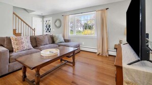 A living room with a sectional sofa, a wooden coffee table, a television, and a large window with curtains. - Downtown Mystic House w/ Yard, Pet-Friendly Getaway (Mystic)
