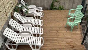 Uma área de piscina com deck de madeira, espreguiçadeiras brancas e uma piscina pequena com escadas de metal.