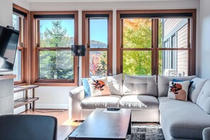 A modern living room with a sofa, a coffee table, and a TV on a stand. There are large windows with a view of trees and a building.