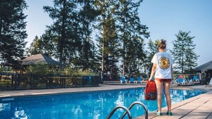 Eine Person steht am Pool, in der Nähe eines Rettungsschwimmer-Schildes, umgeben von Bäumen und einem wolkenlosen Himmel.