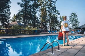Eine Person steht am Pool, in der Nähe eines Rettungsschwimmer-Schildes, umgeben von Bäumen und einem wolkenlosen Himmel.
