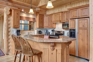A log cabin kitchen with wooden cabinets, granite countertops, and a central island with bar stools. - Le Cerf 40 - Enjoy Nature & Hot tub (Mille-Isles)