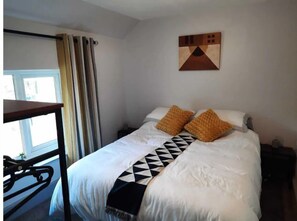 A bedroom with a bed, two pillows, a geometric-patterned blanket, a painting on the wall, a window with curtains, and a bedside table. - Croak Cottage, backing on to Frogmore Creek, part of the Salcombe Estuary (Frogmore, Kingsbridge)