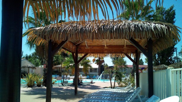 Una zona de piscina a la vora de la platja, amb gandules, una construcció amb teulada de palla i palmeres.