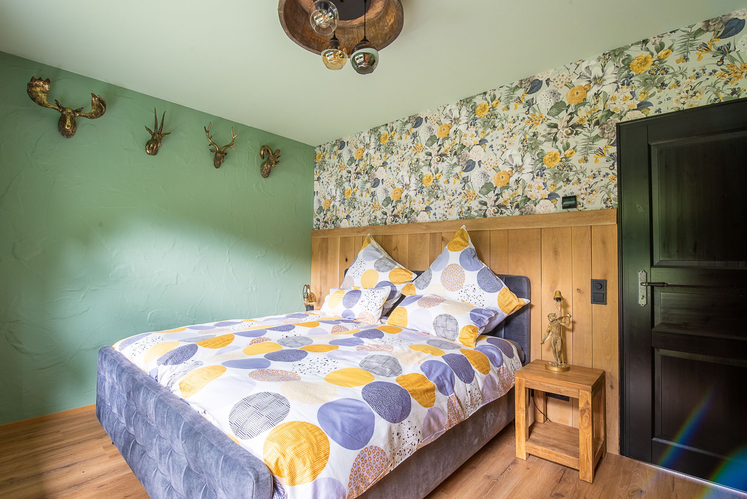 A bedroom with a bed, wooden headboard, floral wallpaper, and a small bedside table with a lamp.