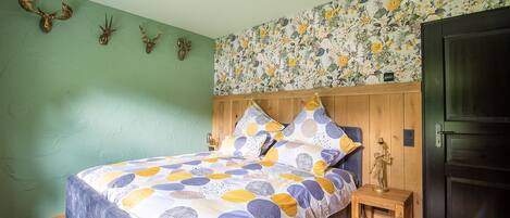 A bedroom with a bed, wooden headboard, floral wallpaper, and a small bedside table with a lamp.