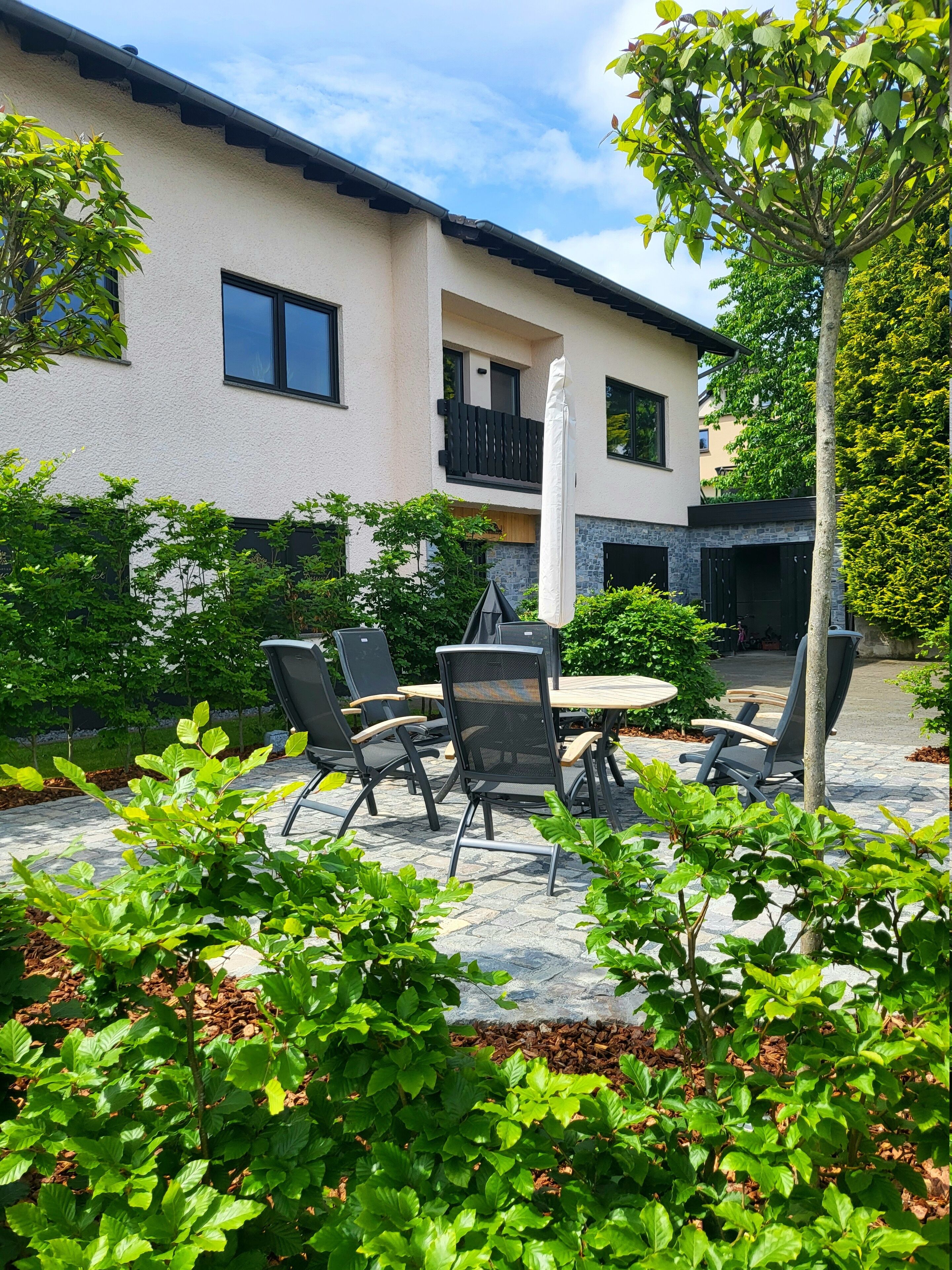 An outdoor patio with a round table and chairs, surrounded by lush greenery and a building with a balcony.