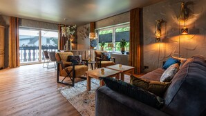 Un salon chaleureux avec un canapé, des fauteuils et une table basse. On y trouve des appliques murales décoratives et une grande fenêtre avec des rideaux.