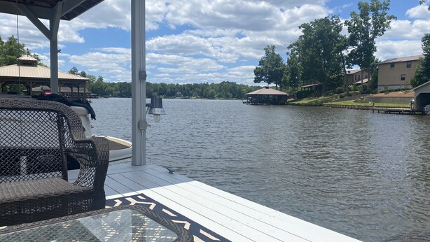 Une jetée aménagée avec des meubles en osier et une table en verre, d’où l’on peut admirer un lac paisible, avec des maisons et un kiosque en arrière-plan.