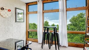 A room with large windows, a clock, a framed picture, a small table, and a bar area with a shelf.