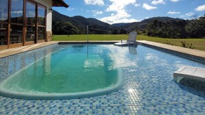A swimming pool with clear blue water, surrounded by a tiled deck. - Chácara Recanto das Princesas (RJ)