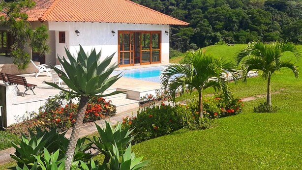A villa with a tiled roof, a swimming pool, and a garden with tropical plants and flowers. - Chácara Recanto das Princesas (RJ)