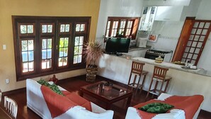 A living room with two white sofas, a wooden coffee table, and a TV. There are windows with wooden frames and a kitchen area in the background. - Chácara Recanto das Princesas (RJ)