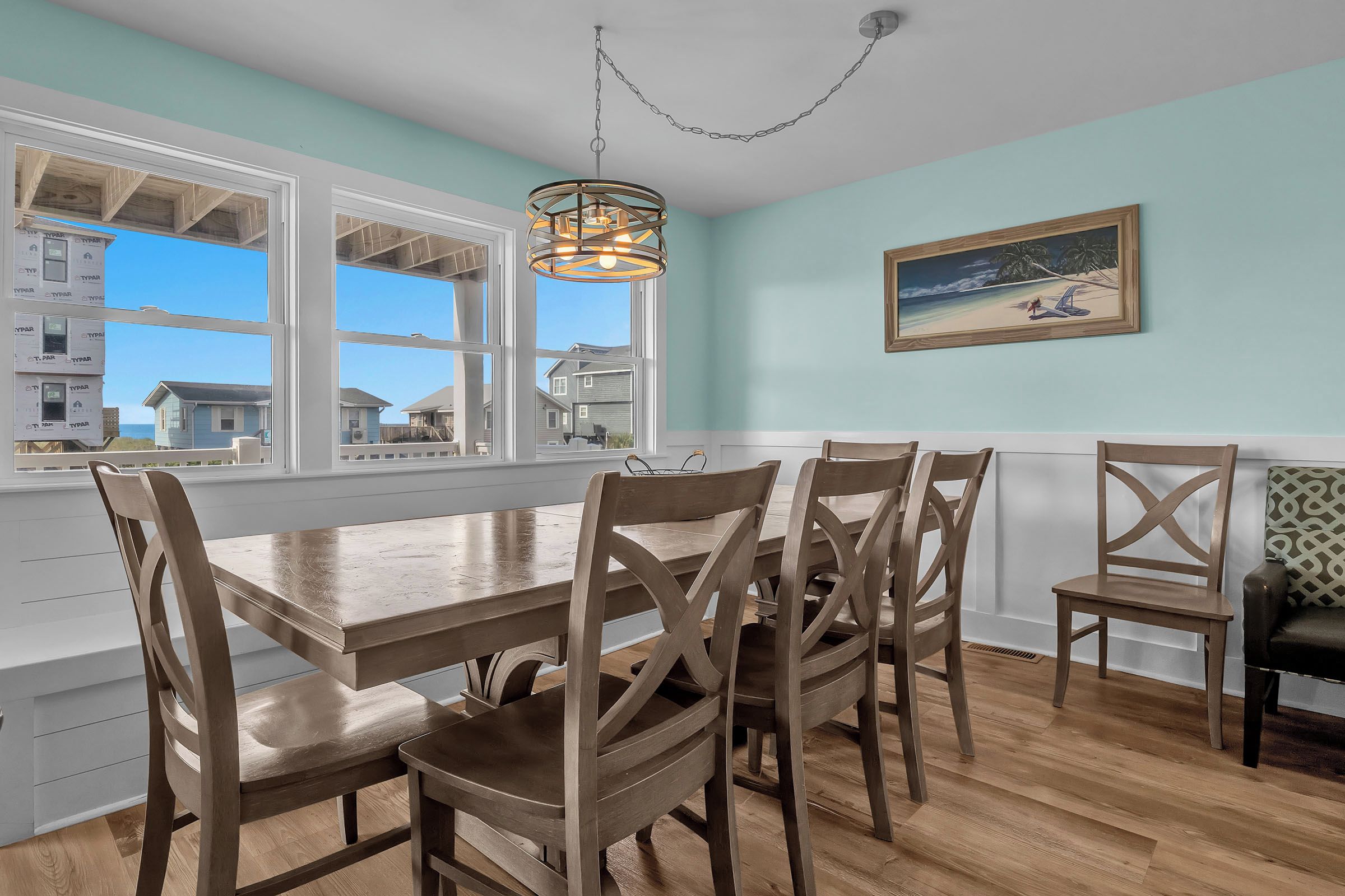 A dining area with a wooden table and chairs, large windows, and a painting on the wall.