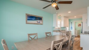 Un coin repas avec une table et des chaises en bois, un ventilateur de plafond et un tableau représentant une scène de plage.