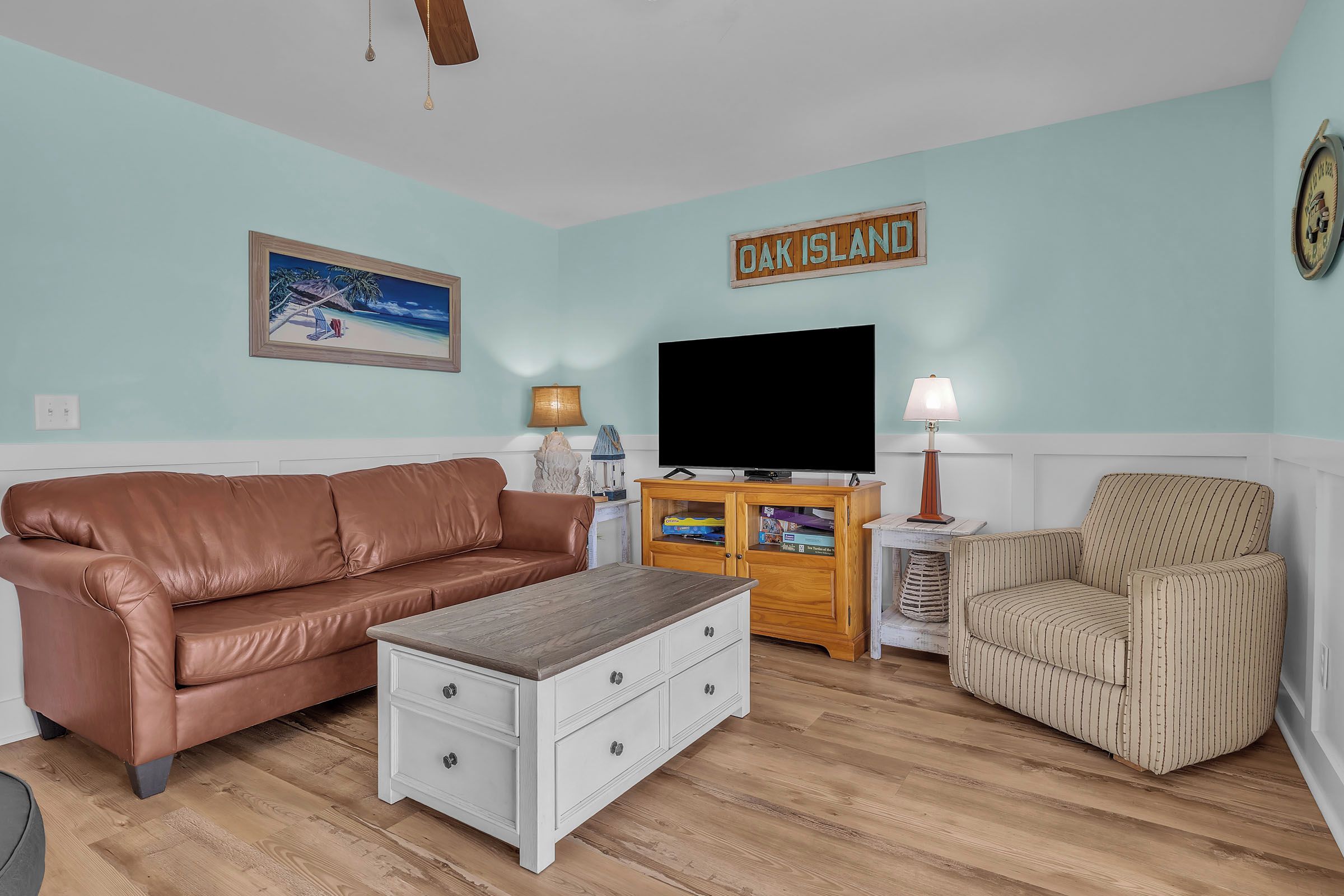 Ein Wohnzimmer mit einem braunen Ledersofa, einem gestreiften Sessel, einem Couchtisch aus Holz, einem Fernseher und einem Schild mit der Aufschrift 'OAK ISLAND'.